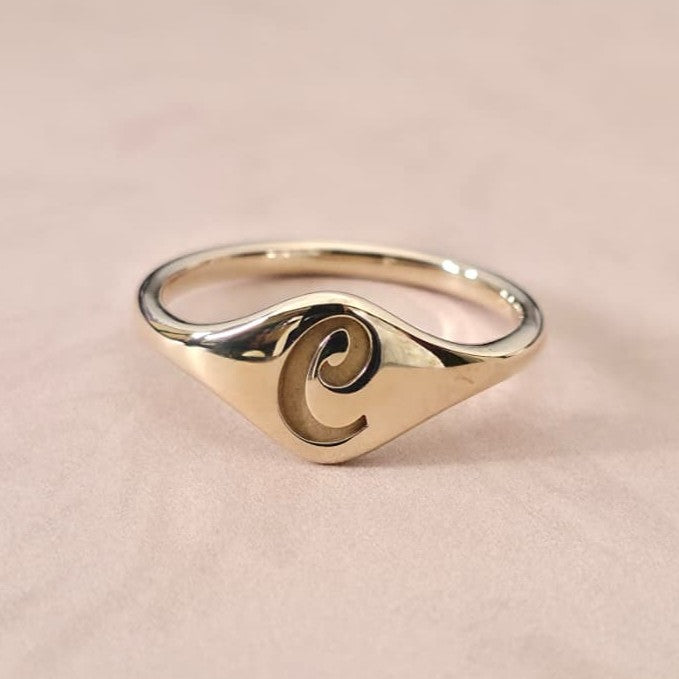 Gold cursive initial signet ring styled for personalized jewelry fashion in soft luxury lighting.