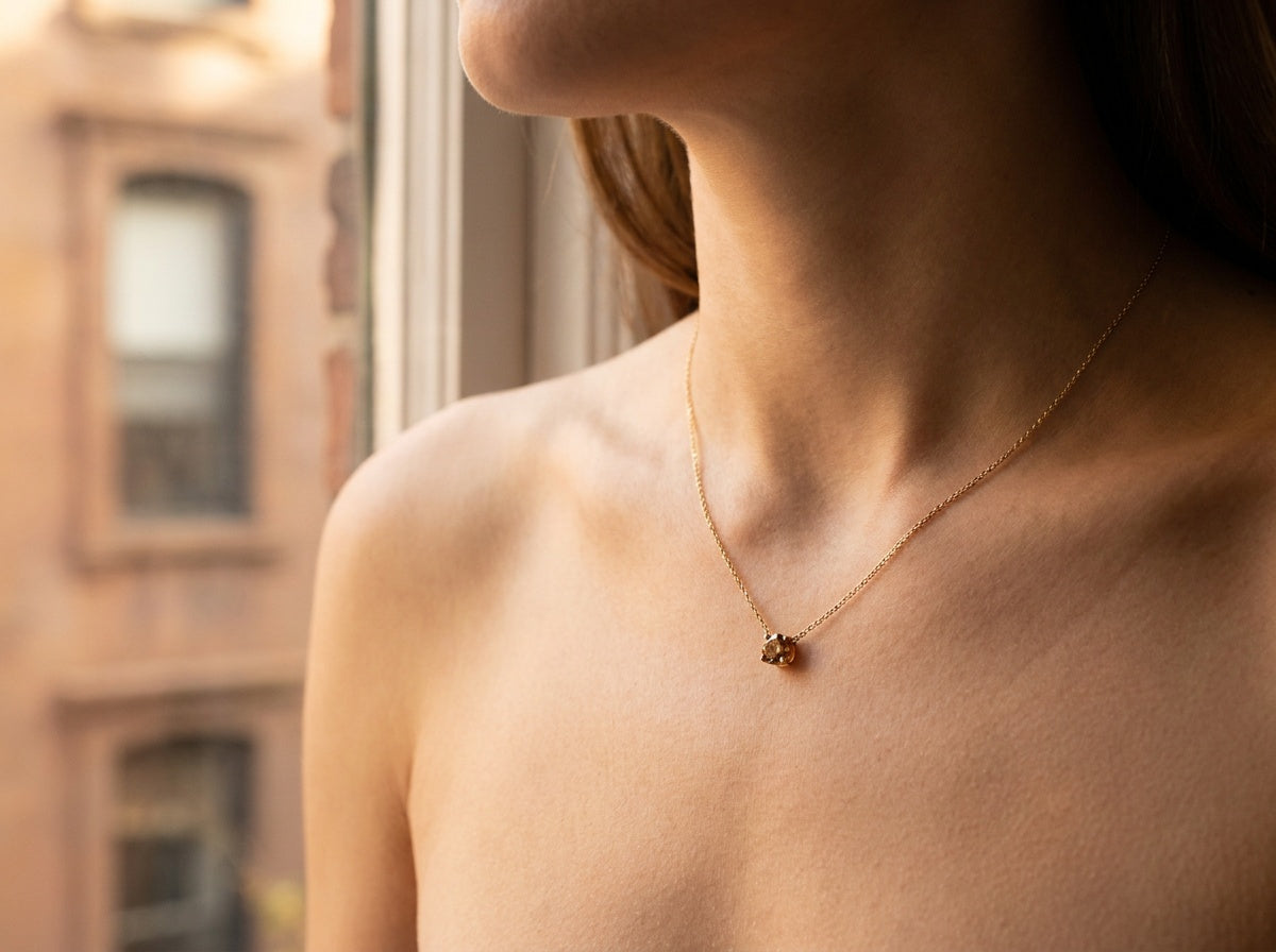 Woman's collarbone wearing a small round cognac diamond solitaire pendant on a fine 14K yellow gold chain, NYC brownstone building visible through window — Brownstone Pendants by Black Pearl of Queens