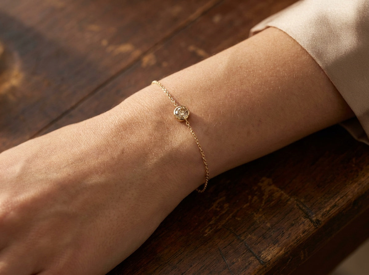 Wrist resting on dark wood wearing a delicate single champagne diamond bezel bracelet on a thin 14K yellow gold chain — Brownstone Bracelets by Black Pearl of Queens