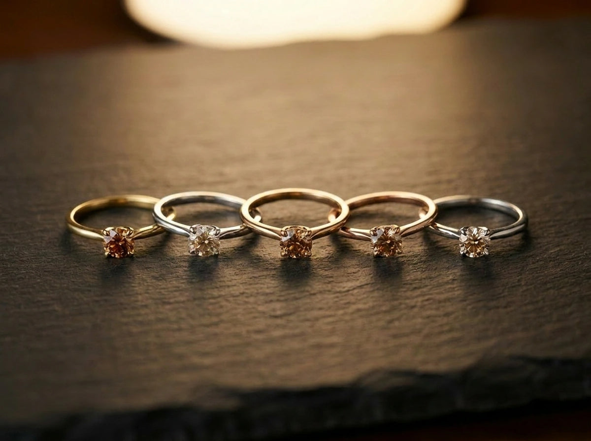 Five cognac diamond solitaire rings in 14K brown gold, yellow gold, rose gold, white gold and platinum arranged on dark slate — Brownstone Collection by Black Pearl of Queens