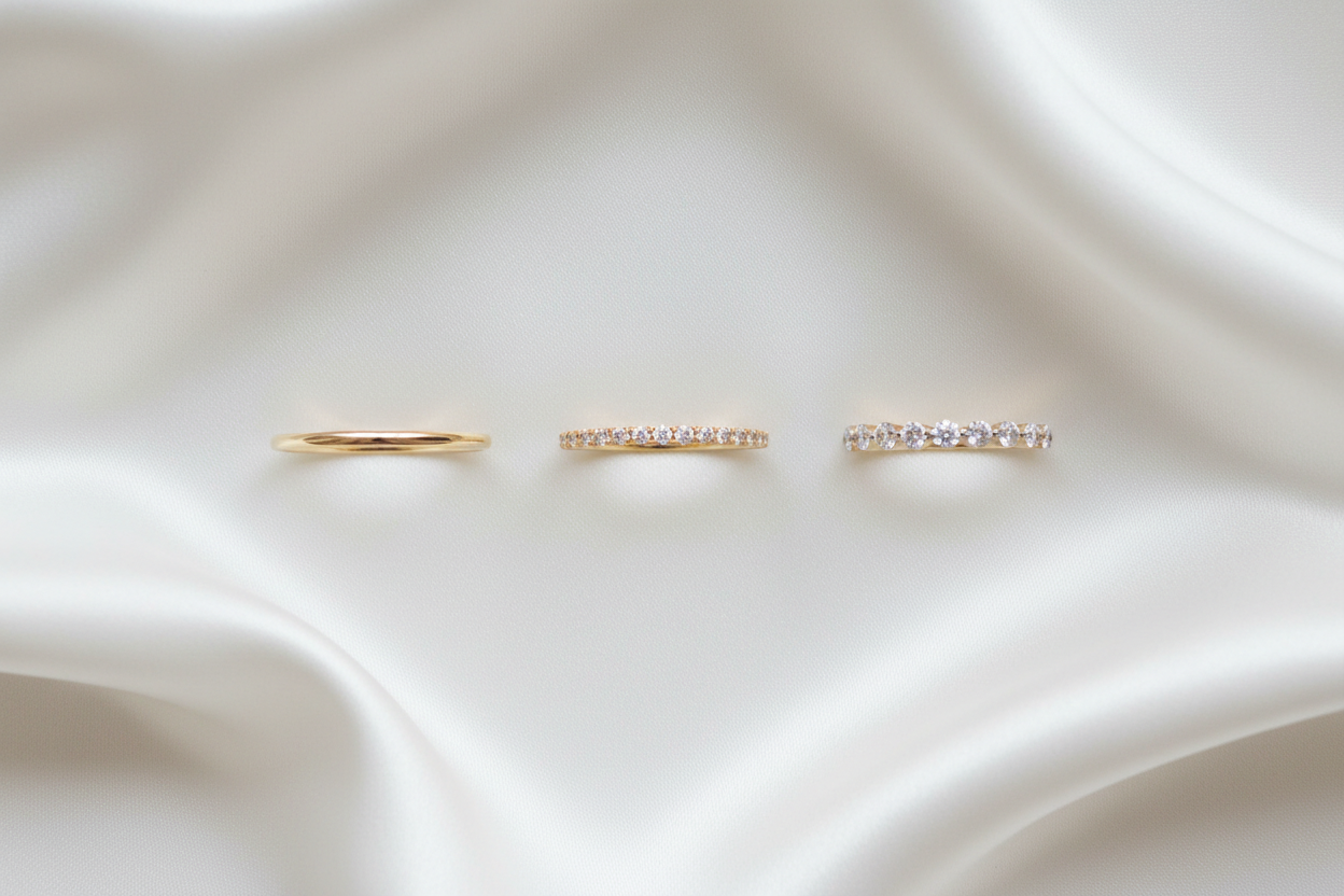 Create a luxury hero image of three 14K yellow gold thin wedding bands arranged side-by-side on a soft ivory bridal background with diffused studio lighting. Left: a very thin 1.5mm plain gold band. Center: a thin pavé diamond band. Right: a thin shared-prong diamond band. Rings should be thin, delicate, evenly spaced, bright, and photographed straight-on with soft shadows. Tiffany & Co. bridal campaign style — elegant, minimal, and soft.