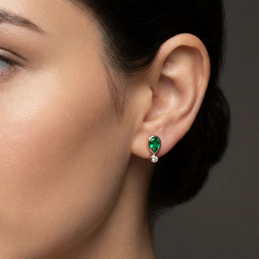 Woman wearing Elyra emerald drop stud earrings with pear shaped green gemstone and diamond detail