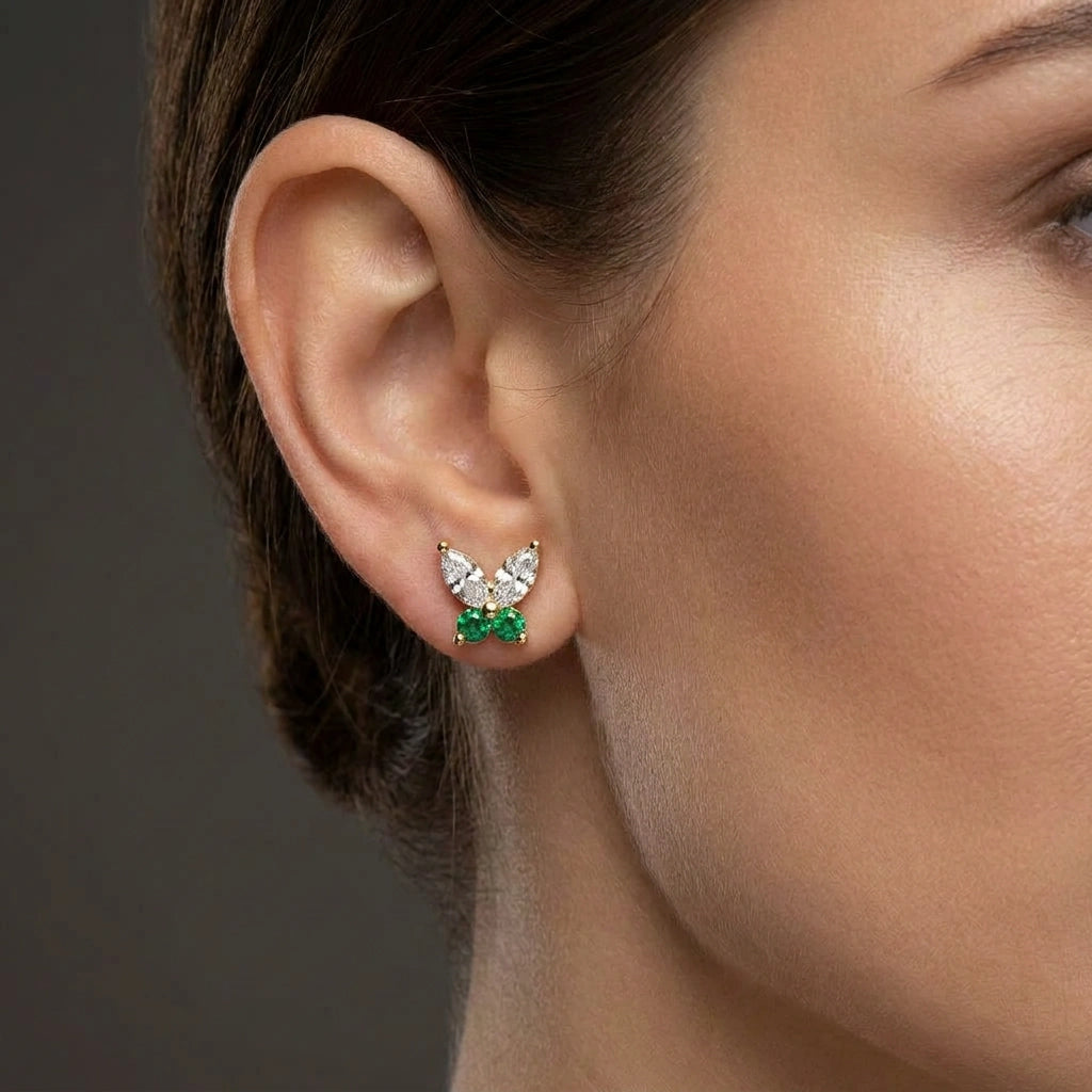 Woman wearing butterfly stud earrings with marquise diamonds and emerald gemstones in yellow gold.