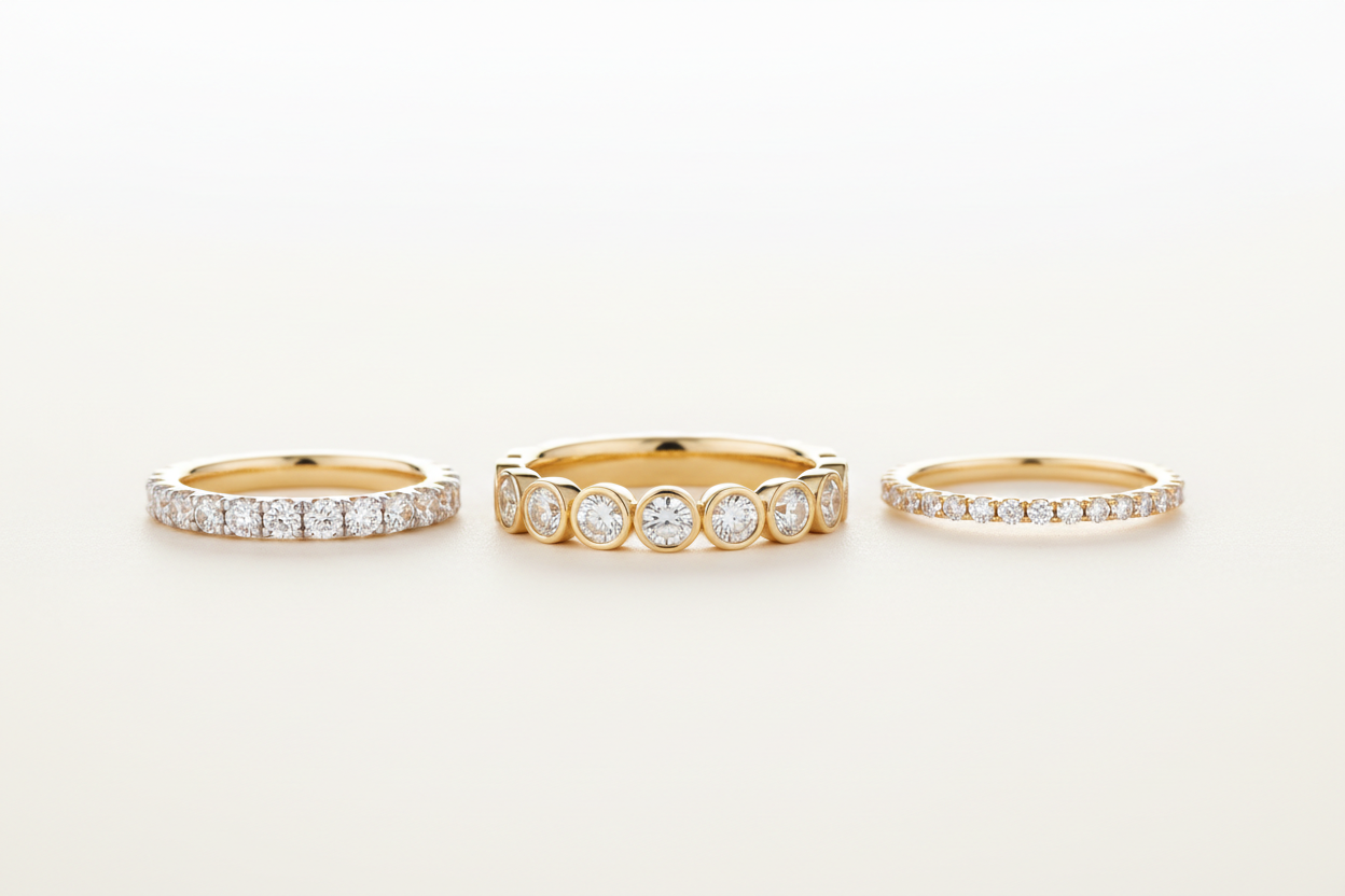 Three 14K gold eternity wedding bands — bezel-set, pavé, and petite styles — displayed side by side on a soft studio background.