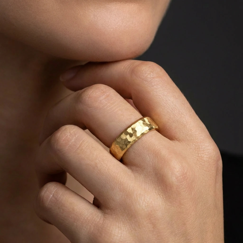 Close-up of a man’s hand wearing a 14K yellow gold hammered wedding band, textured gold ring detail with luxury studio lighting.