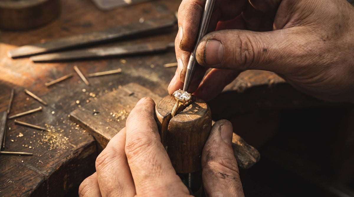 Jeweler setting an oval diamond into the four prongs of an engagement ring at the bench, held in a wooden ring clamp