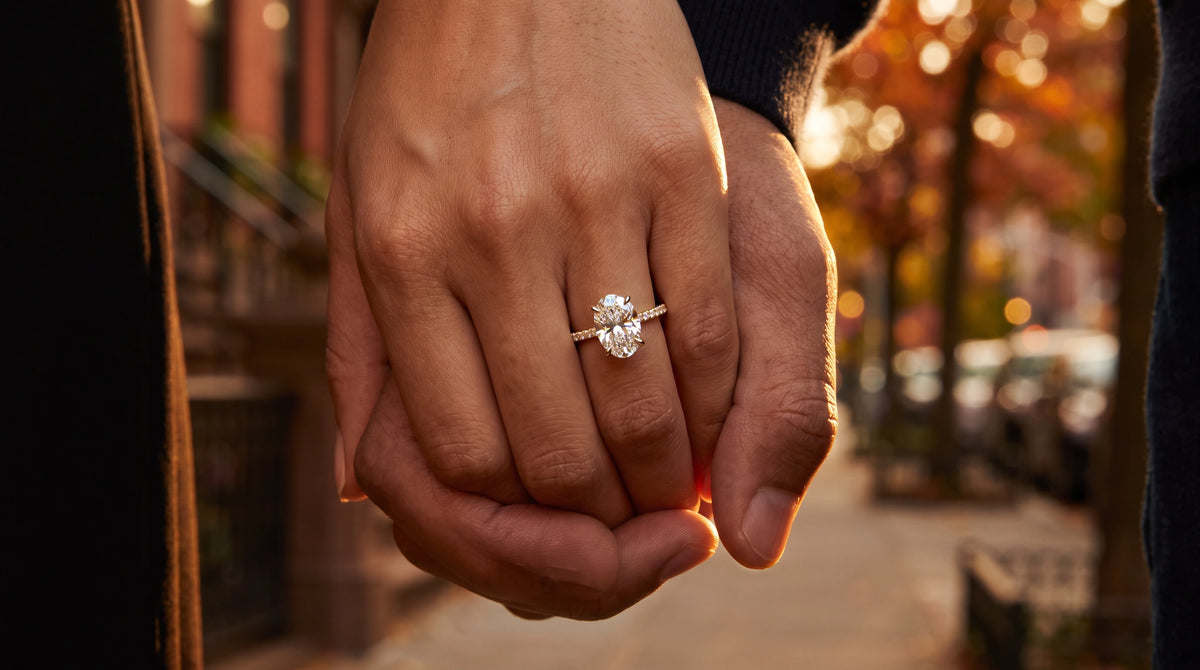 Close-up of an oval pavé Lab-Grown Diamond engagement ring in 14K yellow gold on a woman's hand holding her partner's hand