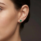 Woman wearing Zirelle emerald halo stud earrings in rose gold with round green emerald center and diamond halo